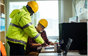 Two men in hard hats working at a desk.